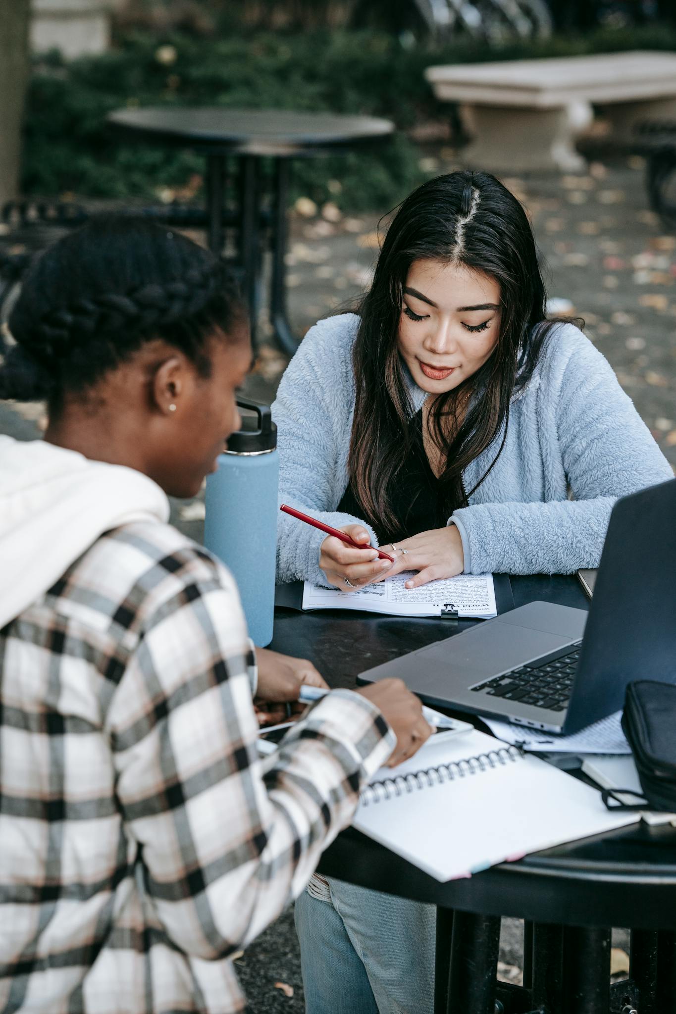 High angle of concentrated young diverse female students writing notes while sitting at table in university campus with laptop and working on research